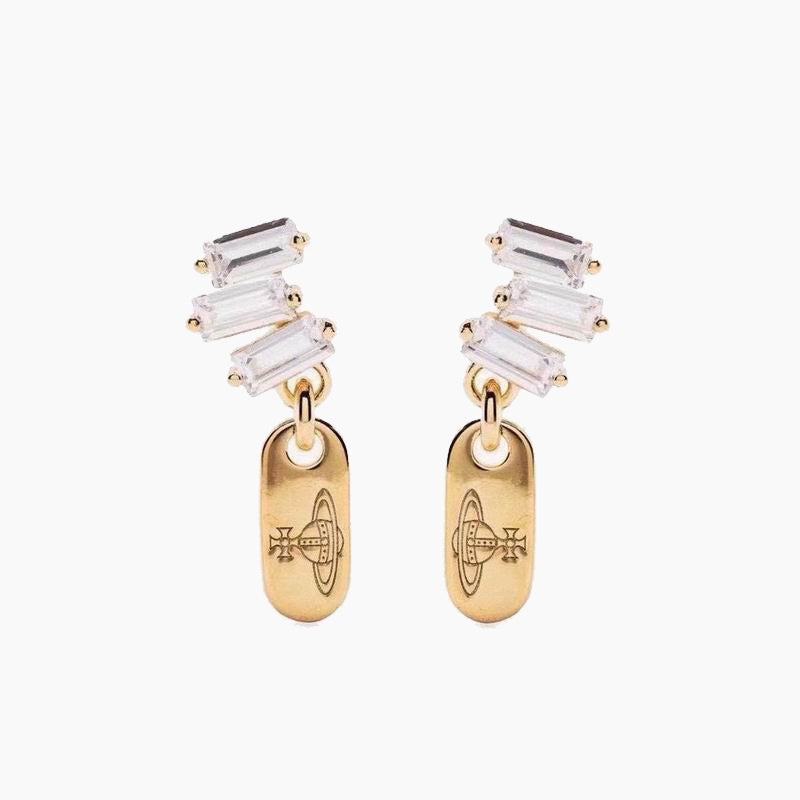 A pair of gold drop earrings featuring a cluster of clear baguette-cut crystals at the top and a polished gold oval pendant below, each engraved with a geometric symbol, set against a plain light gray background.