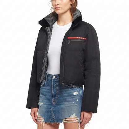 A model wearing a black cropped puffer jacket with a gray patterned inner collar, styled with a white t-shirt and a distressed denim mini skirt against a plain white background.