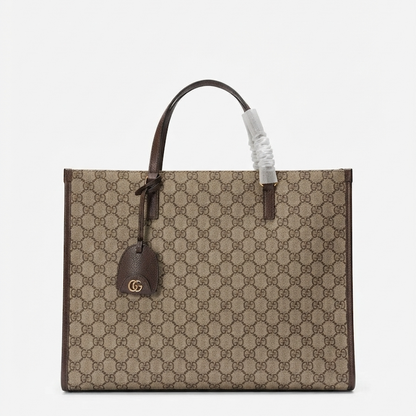 A GG Supreme canvas tote bag in beige and brown, featuring dark brown leather handles and trim, a hanging leather tag with a gold-tone GG logo, and a protective plastic wrap on one handle.