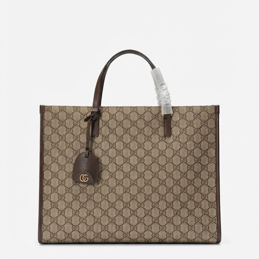 A GG Supreme canvas tote bag in beige and brown, featuring dark brown leather handles and trim, a hanging leather tag with a gold-tone GG logo, and a protective plastic wrap on one handle.