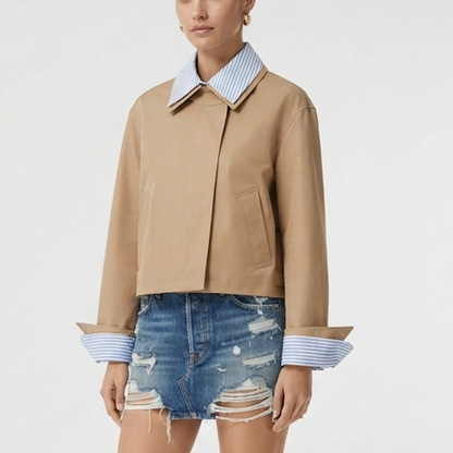 A model wearing a cropped tan jacket with a blue and white striped collar and striped cuffs, paired with distressed denim shorts, against a plain white background.