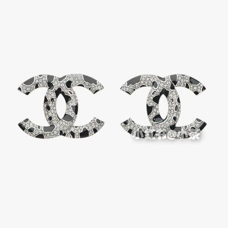 A pair of  logo stud earrings featuring a black and white abstract pattern with sparkling rhinestone-like details, set against a plain light gray background.