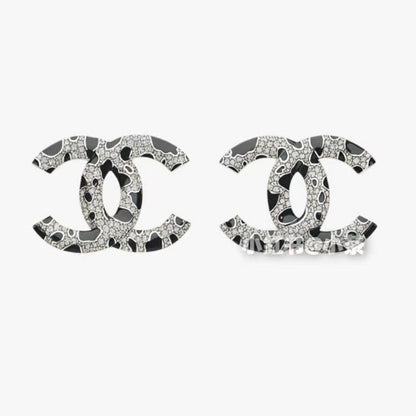 A pair of  logo stud earrings featuring a black and white abstract pattern with sparkling rhinestone-like details, set against a plain light gray background.