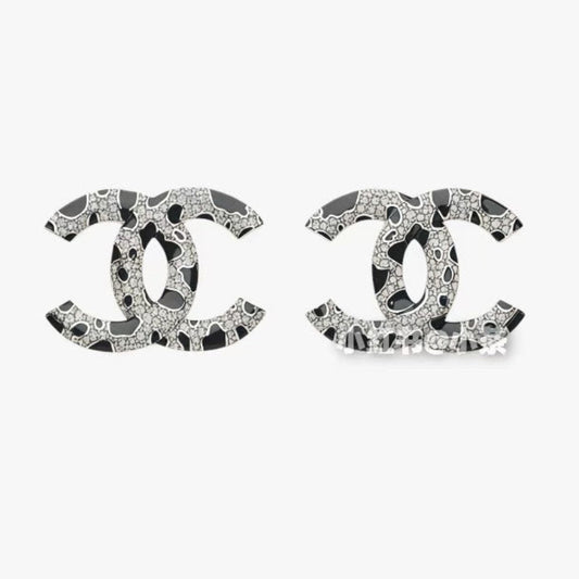 A pair of  logo stud earrings featuring a black and white abstract pattern with sparkling rhinestone-like details, set against a plain light gray background.