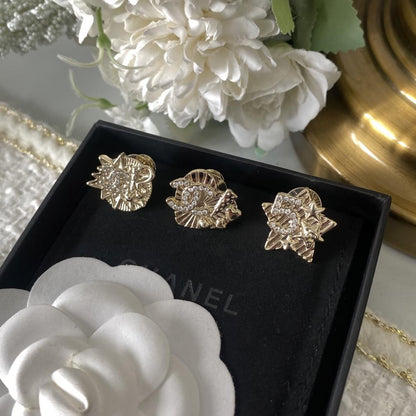 Three gold-toned floral stud earrings displayed in a black box, with white flowers and a gold surface in the background.