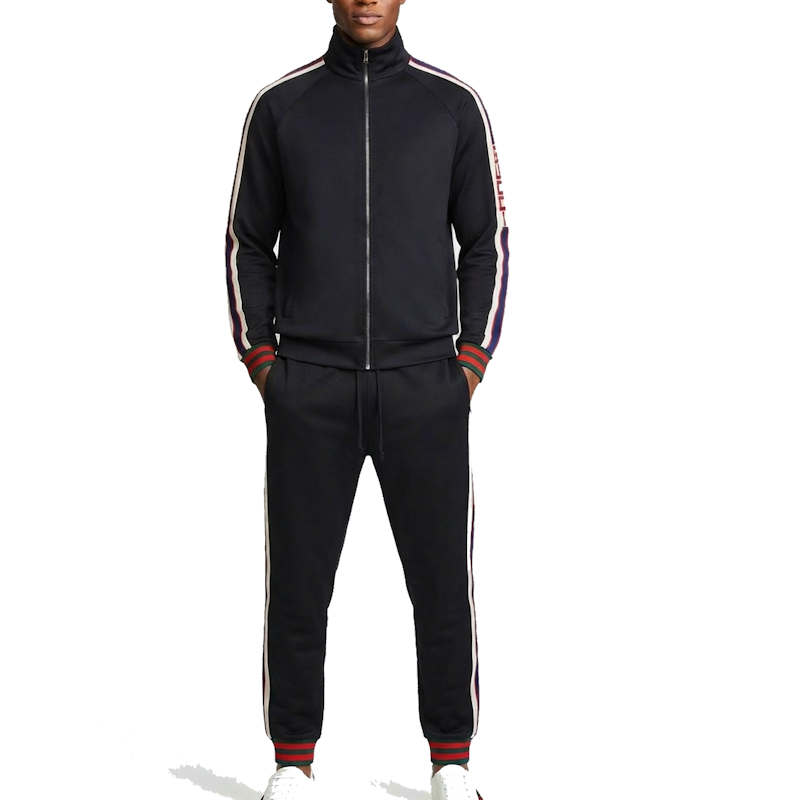 A man wearing a black track suit with white and red stripes down the sleeves and legs, paired with white sneakers, shot against a plain white background.
