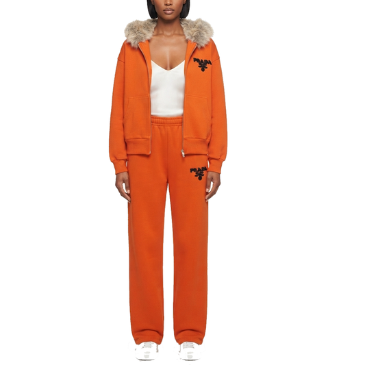 A model wearing a matching orange tracksuit with a faux-fur trimmed hood, featuring a zip-up jacket and matching sweatpants with black embroidered logos, styled with a white top and white sneakers against a plain white background.