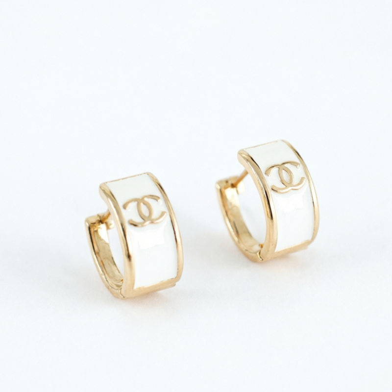 A pair of gold huggie hoop earrings with a white enamel center and the interlocking CC logo, set against a plain white background.