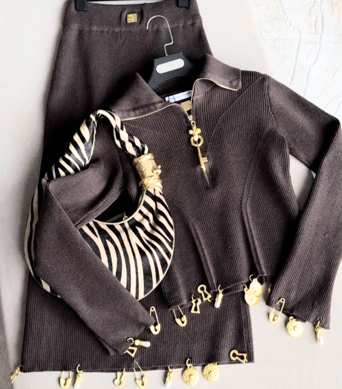 A dark brown ribbed knit long-sleeve top with gold-tone keyhole and safety-pin embellishments, shown folded with a black-and-white striped shoulder bag on a hanger against a light gray background.