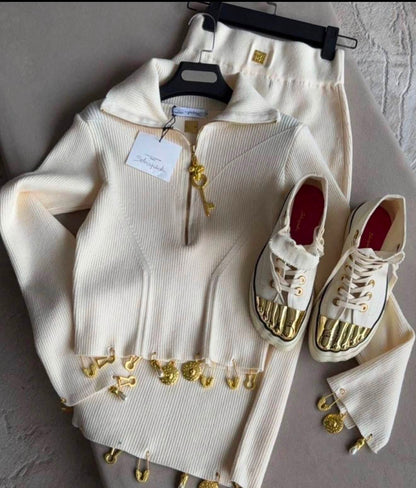 A cream ribbed knit zip-up hoodie with gold-tone hardware and dangling gold charms, shown with matching cream pants on hangers and a pair of cream high-top sneakers with metallic gold toe caps, laid out on a gray surface.