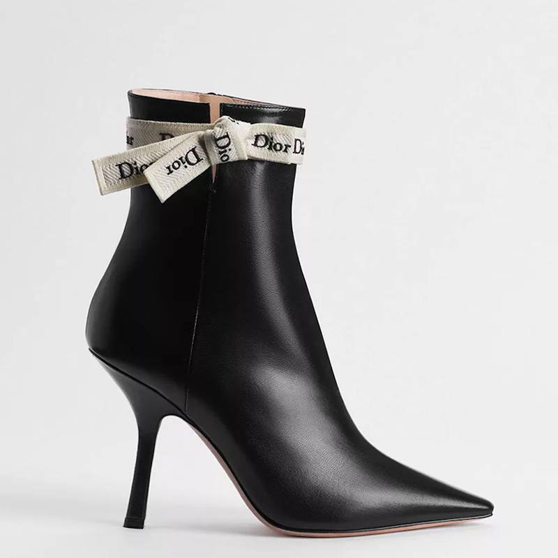 A black leather pointed-toe ankle boot with a slim stiletto heel and a ribbon bow tied around the collar, shown on a light gray background.