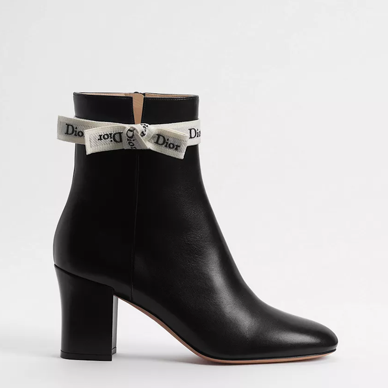 A black leather ankle boot with a block heel, featuring a decorative white ribbon bow with  printed on it around the collar, shown against a plain light gray background.
