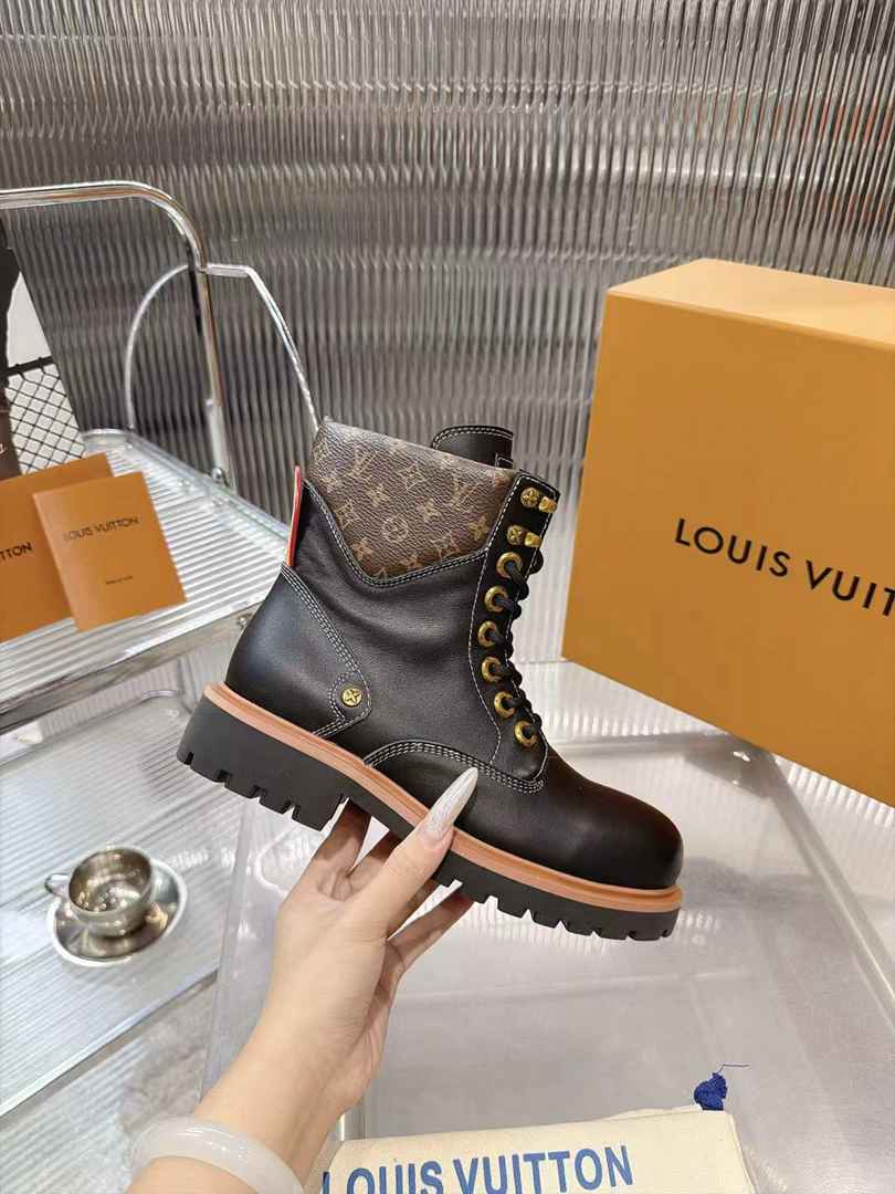 A hand holds a black L lace-up boot with a brown monogram patterned collar and a tan sole, with Louis Vuitton boxes visible in the background.