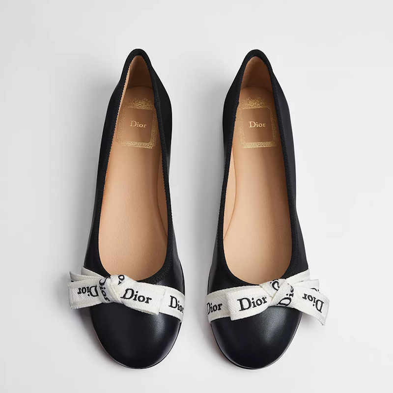 A pair of black leather ballet flats with white ribbon bows printed with the Dior name, shown from a top-down view on a light gray background.