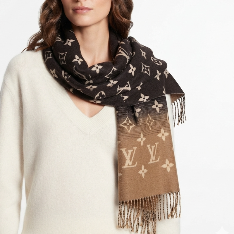 A woman wearing a brown and black monogram scarf with fringed ends, styled over a cream-colored knit sweater against a plain white background.