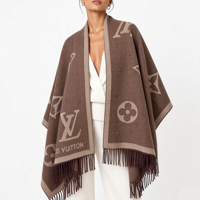 A woman wearing a brown fringed wrap poncho with beige monogram motifs and a white outfit, photographed against a clean white studio background.