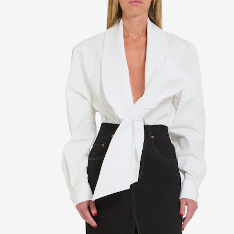 A woman wearing a white wrap-style blazer with a deep V-neck and tied front detail, paired with black jeans, against a plain light background.