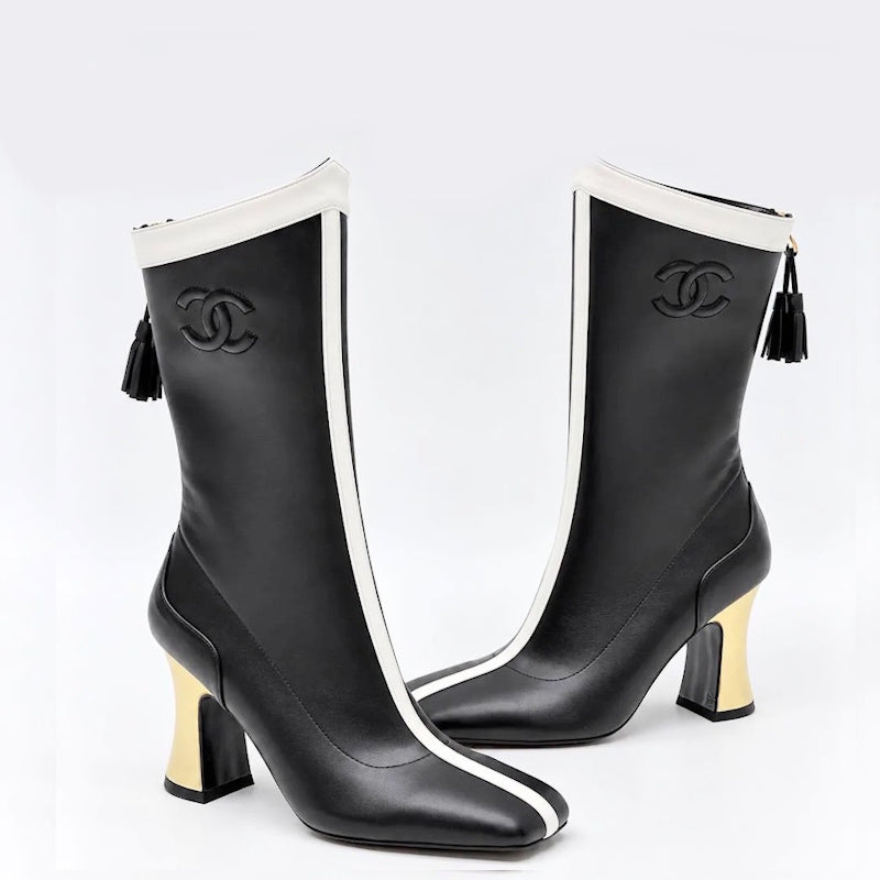 A pair of CC knee-high boots in black leather with white side stripes, black embossed CC logos, black tassel zippers, and gold block heels, shown against a light gray background.