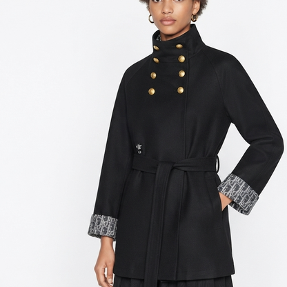 A model wears a black double-breasted coat with gold buttons, a matching waist tie belt, and patterned gray and white cuff linings on a plain white studio background.
