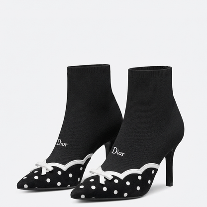A pair of D pointed-toe heeled ankle boots in black knit fabric with white polka dots, white scalloped trim, and white bow details, shown on a light gray background.