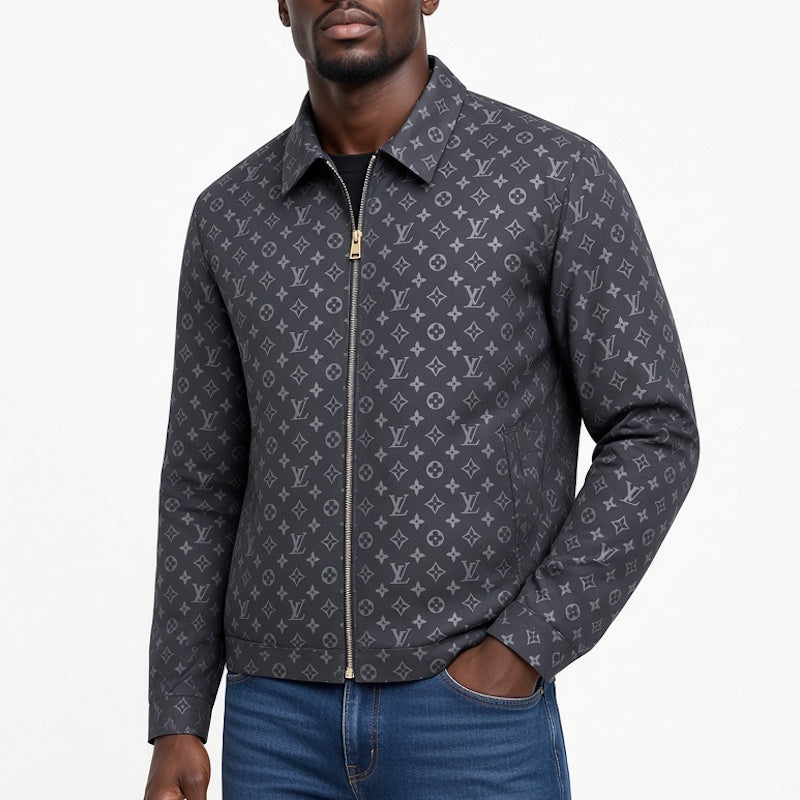 A man wearing a dark gray monogram zip-up jacket over a black shirt and blue jeans, photographed against a plain white studio background.