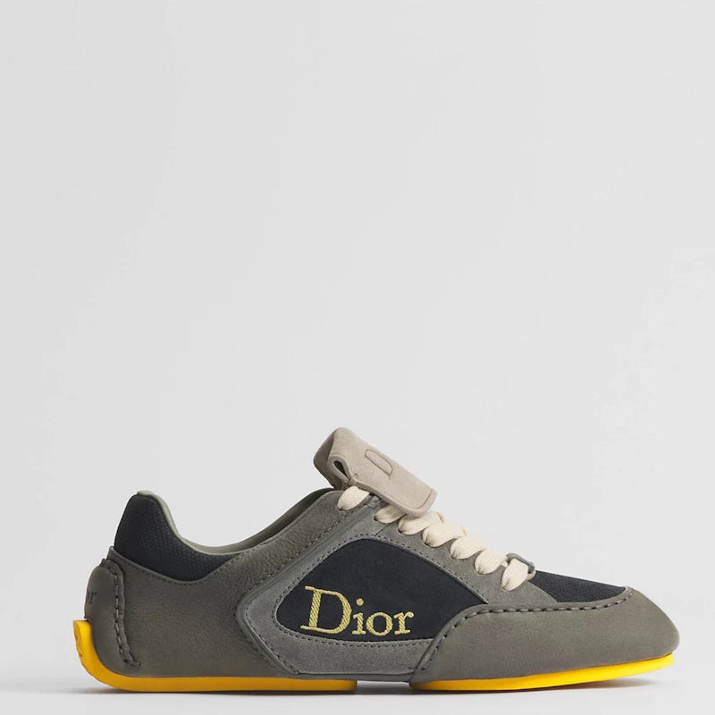 A Di low-top sneaker in olive green suede and black leather, featuring a gold Dior logo on the side, cream laces, and a bright yellow outsole, shown against a light gray background.