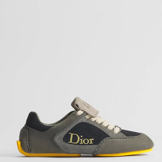 A Di low-top sneaker in olive green suede and black leather, featuring a gold Dior logo on the side, cream laces, and a bright yellow outsole, shown against a light gray background.