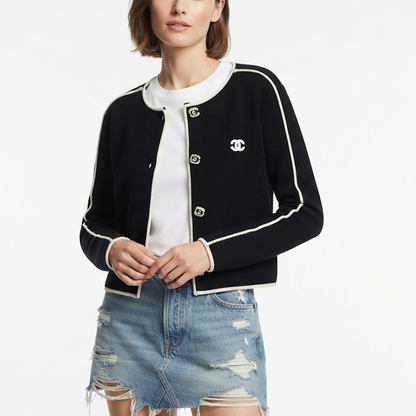 A model wearing a black cardigan with white contrast piping and a white embroidered CC logo on the chest, styled over a white top and a distressed denim mini skirt against a plain white background.