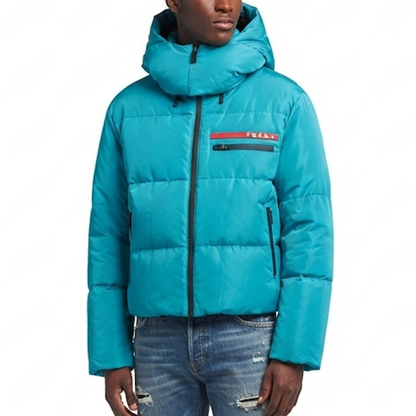 A model wearing a bright turquoise blue puffer jacket with a horizontal quilted design, black zipper, and a red and black logo stripe on the chest. The jacket is paired with distressed blue jeans against a plain white background.