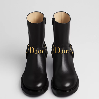 A pair of black leather ankle boots with gold-tone hardware and a prominent  logo strap, photographed from a front-facing angle on a light gray background.