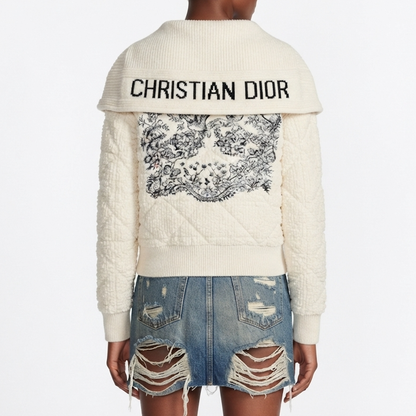 A model shown from the back wearing a cream-colored ribbed knit sweater with embroidered in black across the shoulders and a large black toile-style pattern across the back. The model is also wearing distressed denim shorts against a plain white background.