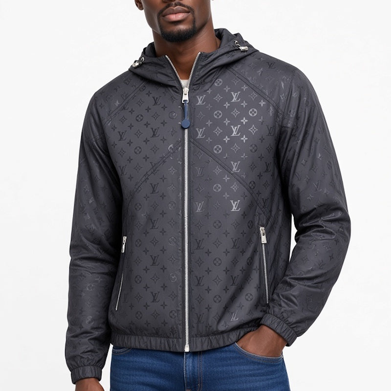 A man wearing a dark gray  hooded jacket with an all-over monogram pattern, featuring a full front zipper and two zippered hand pockets, paired with blue jeans against a plain white background.