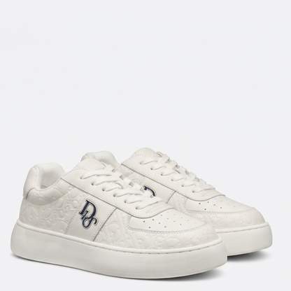 A pair of white low-top sneakers with embossed monogram patterns, featuring a navy blue 'BD' logo on the side and matching white laces, set against a plain light gray background.