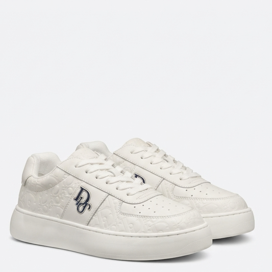 A pair of white low-top sneakers with embossed monogram patterns, featuring a navy blue 'BD' logo on the side and matching white laces, set against a plain light gray background.