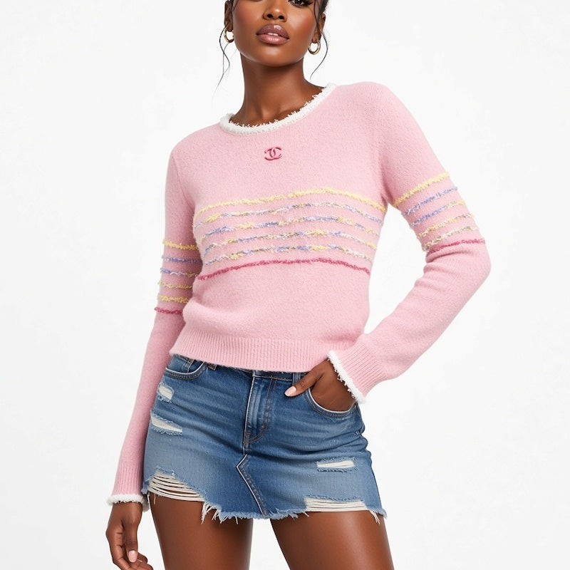 A woman models a light pink knit sweater with colorful striped details across the chest and sleeves, paired with a distressed denim mini skirt against a plain white background.