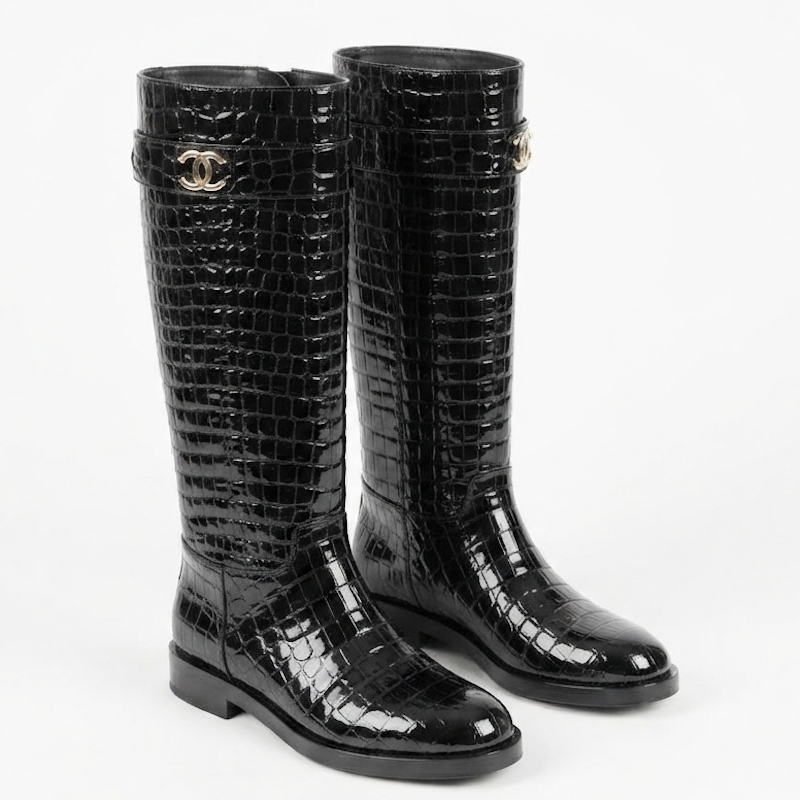 A pair of black knee-high rain boots with a glossy crocodile-embossed texture, featuring a silver-tone interlocking CC logo on the side, characteristic of Chanel.