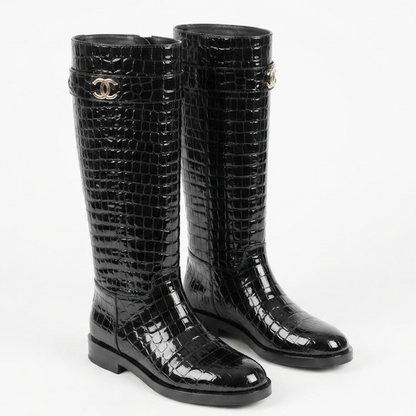 A pair of black knee-high rain boots with a glossy crocodile-embossed texture, featuring a silver-tone interlocking CC logo on the side, characteristic of Chanel.