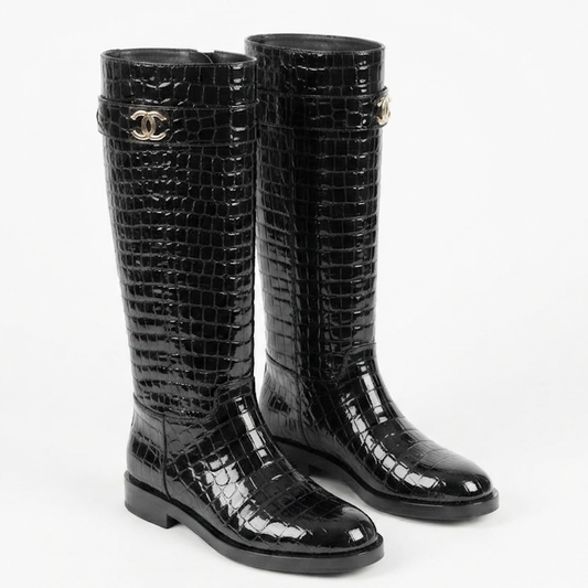 A pair of black knee-high rain boots with a glossy crocodile-embossed texture, featuring a silver-tone interlocking CC logo on the side, characteristic of Chanel.