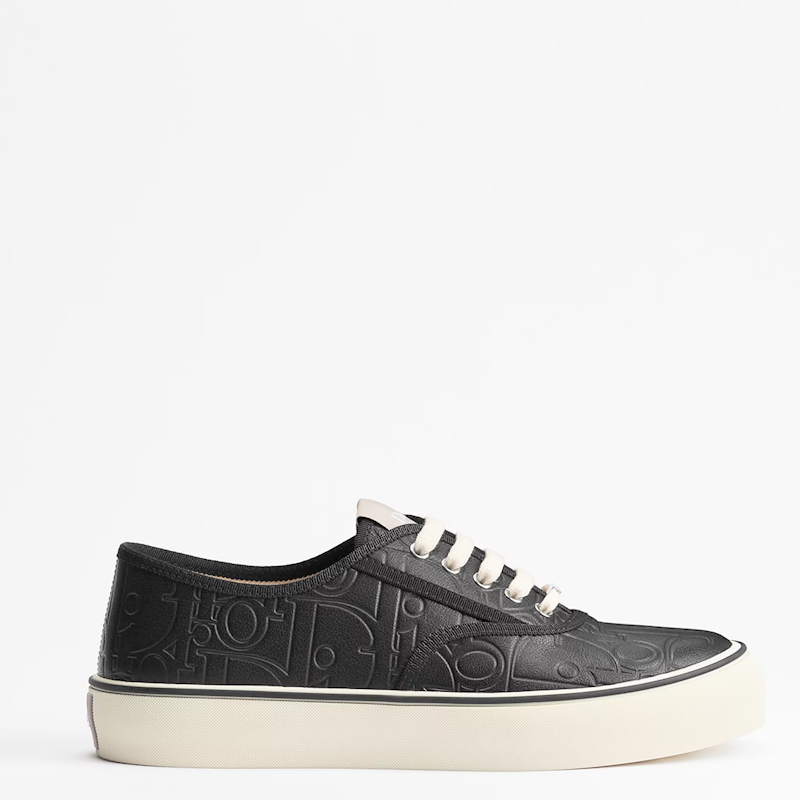 A black leather low-top sneaker with an embossed monogram pattern, white laces, and a thick off-white platform sole, shown from a side profile on a white background.
