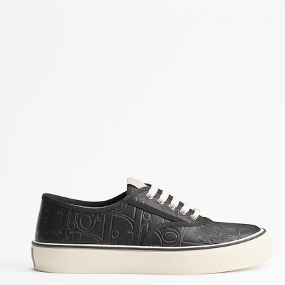 A black leather low-top sneaker with an embossed monogram pattern, white laces, and a thick off-white platform sole, shown from a side profile on a white background.