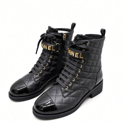A pair of black quilted combat boots with patent leather toes, black laces, and gold-tone hardware. The straps feature the brand name  in gold lettering, and the boots are set against a plain white background.