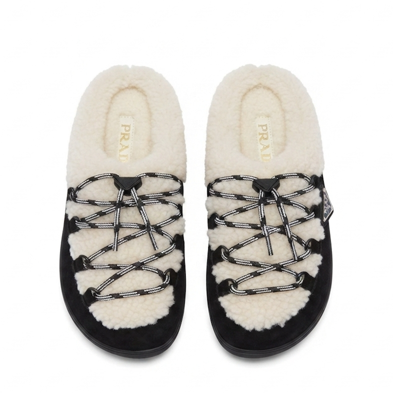 A pair of Prada slip-on slippers featuring a black suede-like base, thick cream shearling lining, and decorative black-and-white patterned laces, shown against a plain white background.