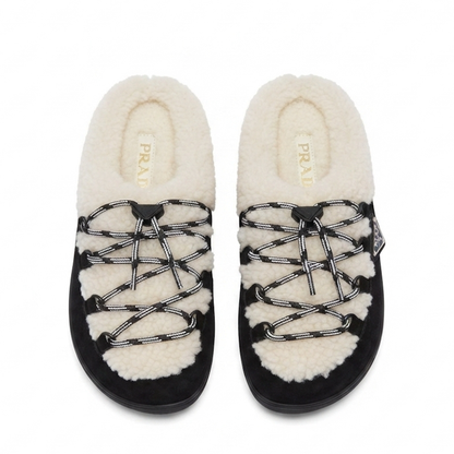 A pair of Prada slip-on slippers featuring a black suede-like base, thick cream shearling lining, and decorative black-and-white patterned laces, shown against a plain white background.