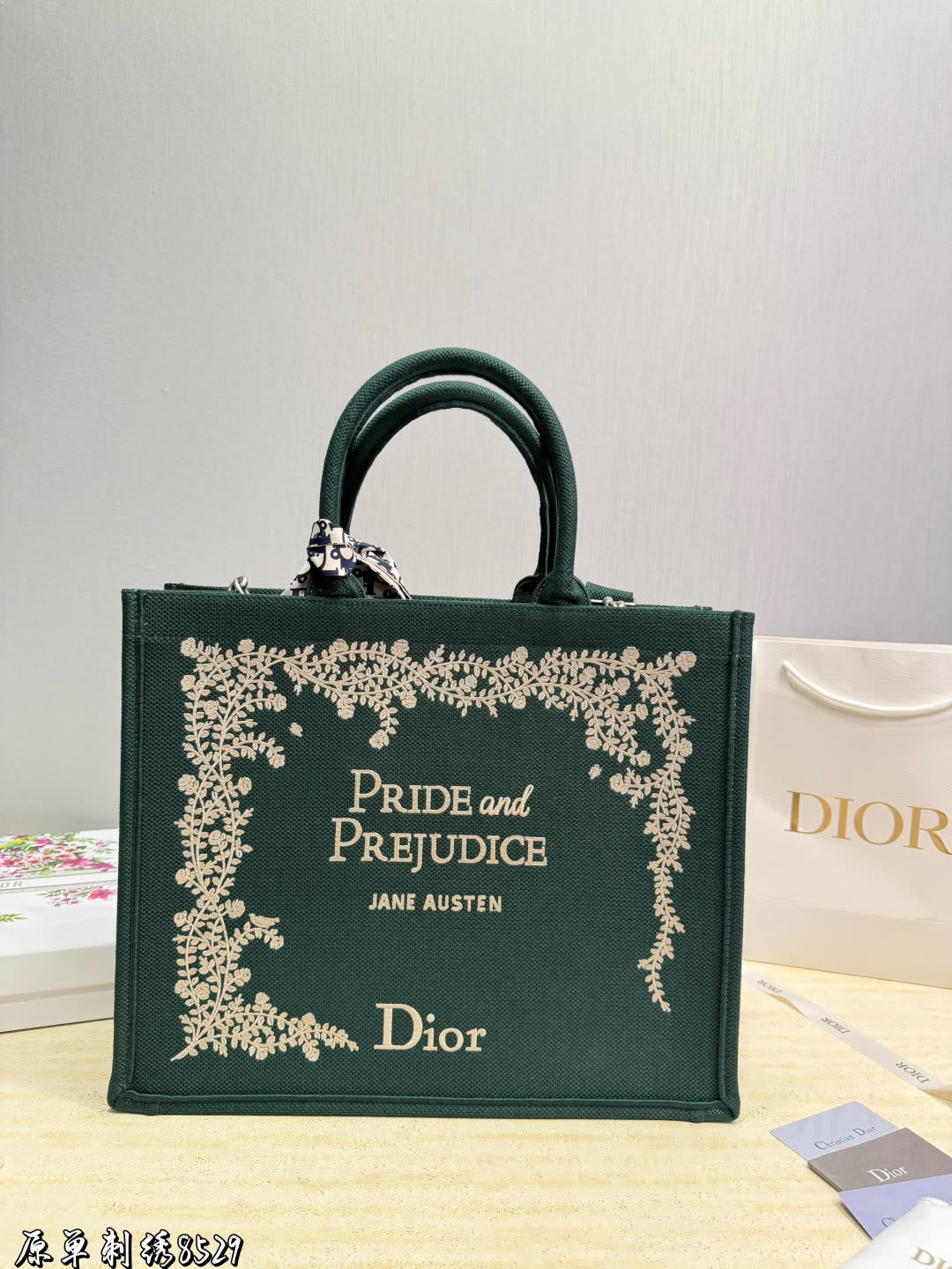 A dark green  tote bag featuring gold foil floral borders and the text 'PRIDE and PREJUDICE JANE AUSTEN' on the front, displayed on a light wooden surface with a Dior shopping bag in the background.