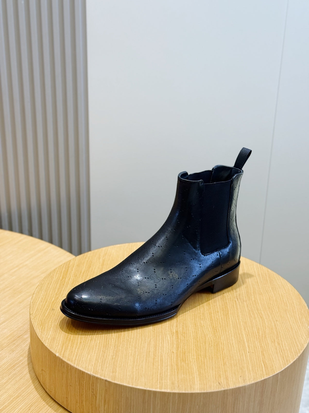 A single dark navy blue leather Chelsea boot with perforated detailing on the toe, displayed on a round light wood pedestal.