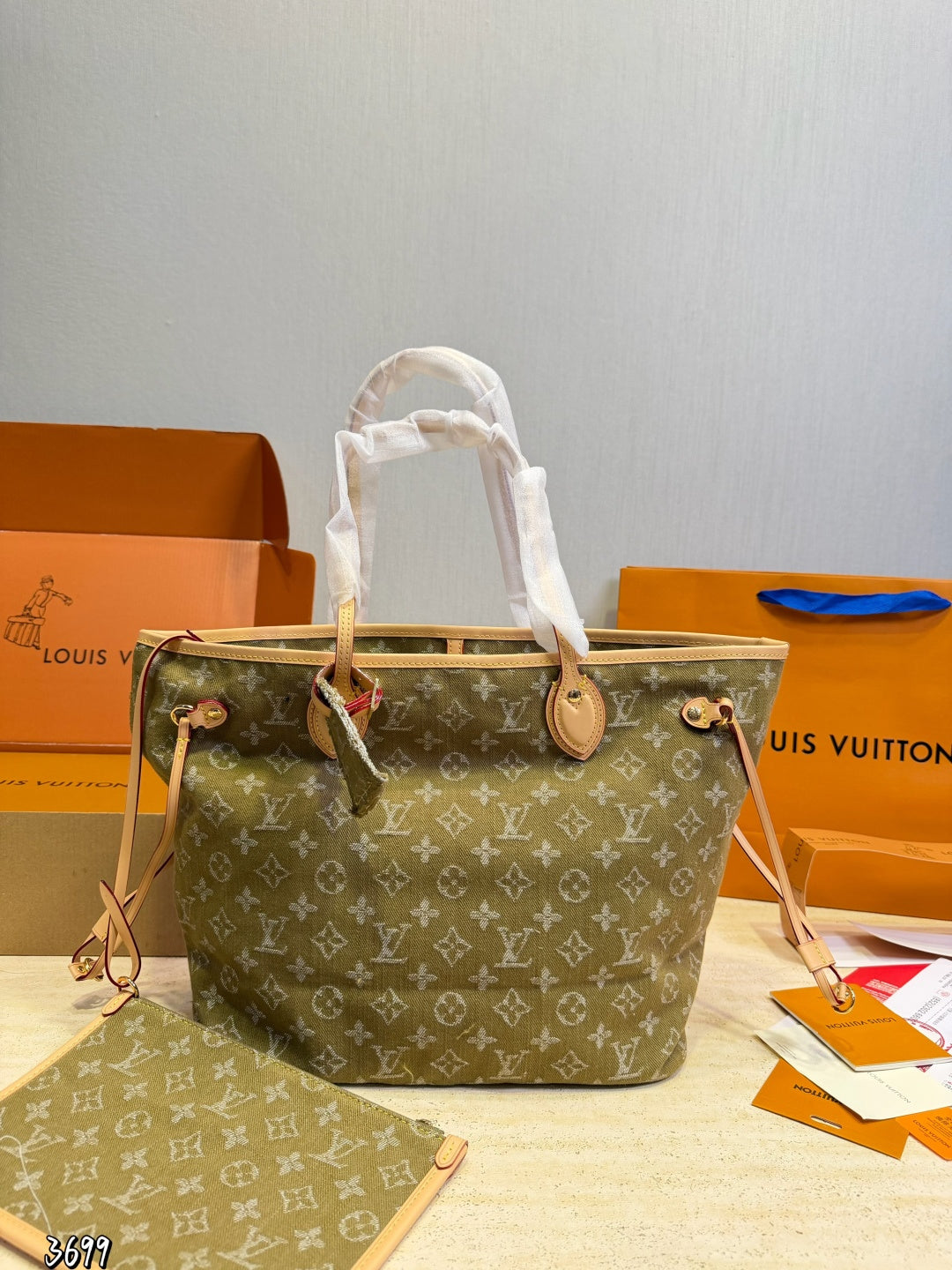 monogram canvas tote bag in olive green with beige leather trim and white protective paper wrapping on the handles, displayed with orange Louis Vuitton boxes and shopping bags in the background.