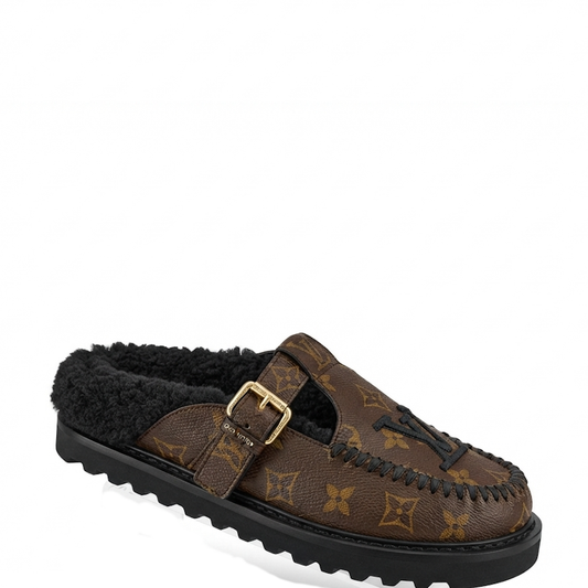 monogram canvas slip-on moccasin-style shoe with black shearling lining, a gold-tone buckle strap, and a black lug sole, shown on a white background.