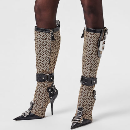 A pair of knee-high pointed-toe stiletto boots with a beige and black monogram pattern, black faux-leather straps, and silver hardware, shown on a model's legs against a light gray background.