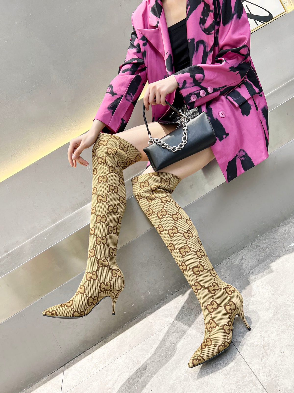 A model wearing beige over-the-knee boots with a brown GG monogram pattern, paired with a bright pink and black patterned blazer and a black handbag.