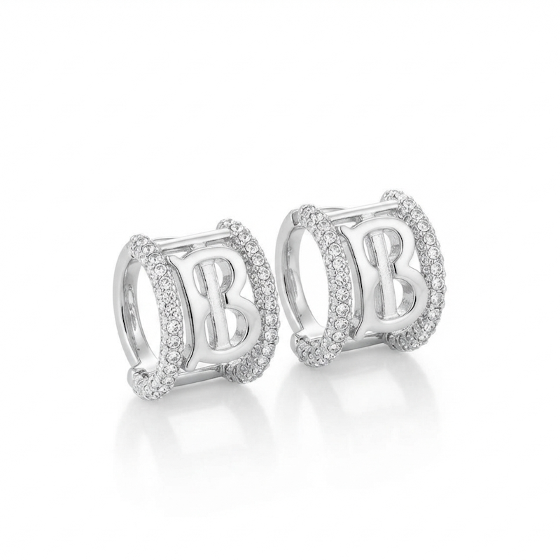 A pair of silver-toned huggie hoop earrings featuring a central letter 'B' surrounded by pavé-set clear stones, shown on a clean white background with a soft reflection.